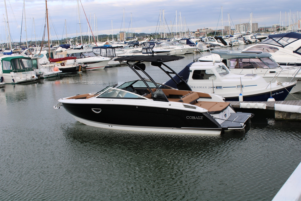 Cobalt boat docked at a marina surrounded by various yachts and boats under a cloudy sky, reflecting on the water.